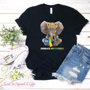 May include: Black t-shirt with a cartoon elephant wearing a flower crown and a blue and yellow ribbon. The text "Embrace Differences" is printed below the elephant.
