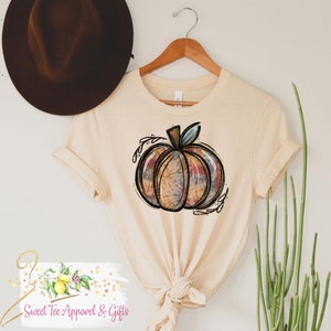 May include: A beige t-shirt with a tie-dye pumpkin graphic. The pumpkin is outlined in black and has a green stem. The graphic is surrounded by small black leaves.