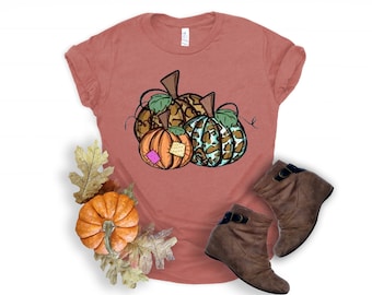May include: A rust-colored t-shirt with a graphic of three pumpkins. The pumpkins are brown, orange, and teal with a leopard print pattern.