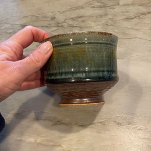 May include: A small, green and brown ceramic bowl with a textured base. The bowl has a glazed finish and a slightly flared rim.