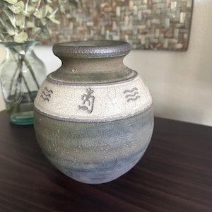 May include: A handcrafted ceramic vase with a dark brown rim and a textured, crackled white band. The vase features a blue and green body with embossed symbols and wave designs. A decorative piece for home decor.
