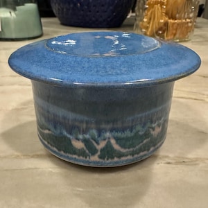 May include: A blue ceramic butter dish with a lid. The dish has a light blue glaze with a darker blue rim and a green and pink design on the bottom.