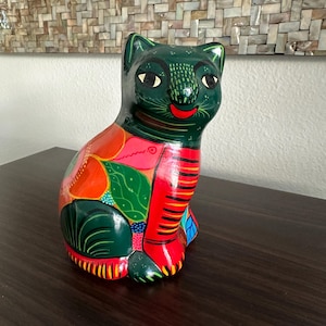 May include: A hand-painted ceramic cat figurine. The cat is primarily green with colorful accents of red, orange, and blue. The cat has a smiling red mouth and is sitting upright.