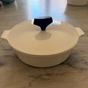 May include: A round, off-white casserole dish with a matching lid and two side handles. The lid features a prominent, black, T-shaped knob centered on top. The dish has a smooth, solid-colored finish and a low-profile, wide-rimmed design.