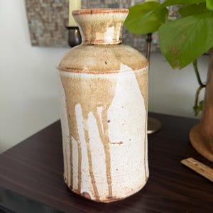 May include: A handmade ceramic vase with a cylindrical body and a flared neck. The vase is primarily off-white with brown drips and accents. The vase has a rustic, textured appearance, suggesting a pottery style.