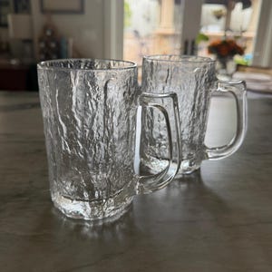 May include: Two clear glass mugs featuring a textured, bark-like exterior surface. Each mug has a smooth, curved handle and a cylindrical body with a thick base. The glass is transparent and reflects light across the uneven, rugged pattern.