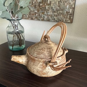 May include: A handcrafted ceramic teapot with a textured, speckled brown finish. It features a woven handle and a spout. A glass vase with greenery is to the left, and a woven wall hanging is in the background.