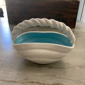May include: White ceramic bowl with a blue interior and a rope-like handle. The bowl has a wavy, organic shape.