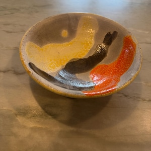 May include: A handmade ceramic bowl with a unique design. The bowl features a gray exterior with yellow, orange, and black abstract shapes on the inside. The rim is yellow, and the bowl is sitting on a marble surface.