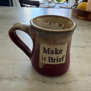 May include: A ceramic mug with a deep red base and a tan drip glaze around the rim and handle. A rectangular plaque on the front features the black text "Make it Brief" in a serif font. The interior is off-white with a tan drip pattern at the top.