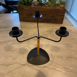 May include: A black metal candle holder with three arms and a woven brown base. The candle holder is on a white marble surface.