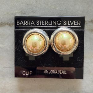 May include: A pair of clip-on earrings on a black card. The earrings have a silver-tone metal frame with a gold-colored center. The card reads "BARRA STERLING SILVER" and "MALLORCA PEARL."