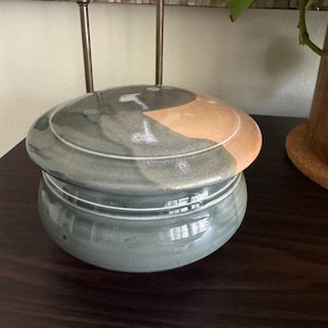 May include: A round ceramic lidded jar with a glossy finish. The base is a muted sage green, while the lid features a blend of sage green and a warm, earthy peach tone. The container has a smooth, curved silhouette with a distinct horizontal rim.