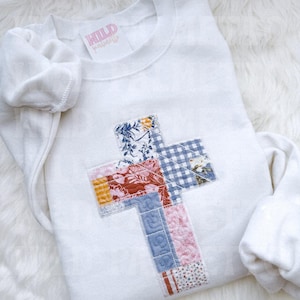 May include: A white sweatshirt with a patchwork cross design. The cross is made of various patterned fabrics, including floral, gingham, and striped patterns. The sweatshirt has a crew neck and long sleeves. The brand name "WILD PAISLEY" is printed on a pink label.