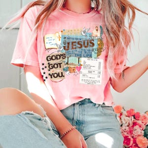 May include: A light pink t-shirt with a graphic design featuring the text "DO YOU KNOW JESUS THE WAY THE TRUTH THE LIFE" and "GOD'S GOT YOU." The design includes doves, a cross, and a depiction of Jesus. The shirt is short-sleeved.