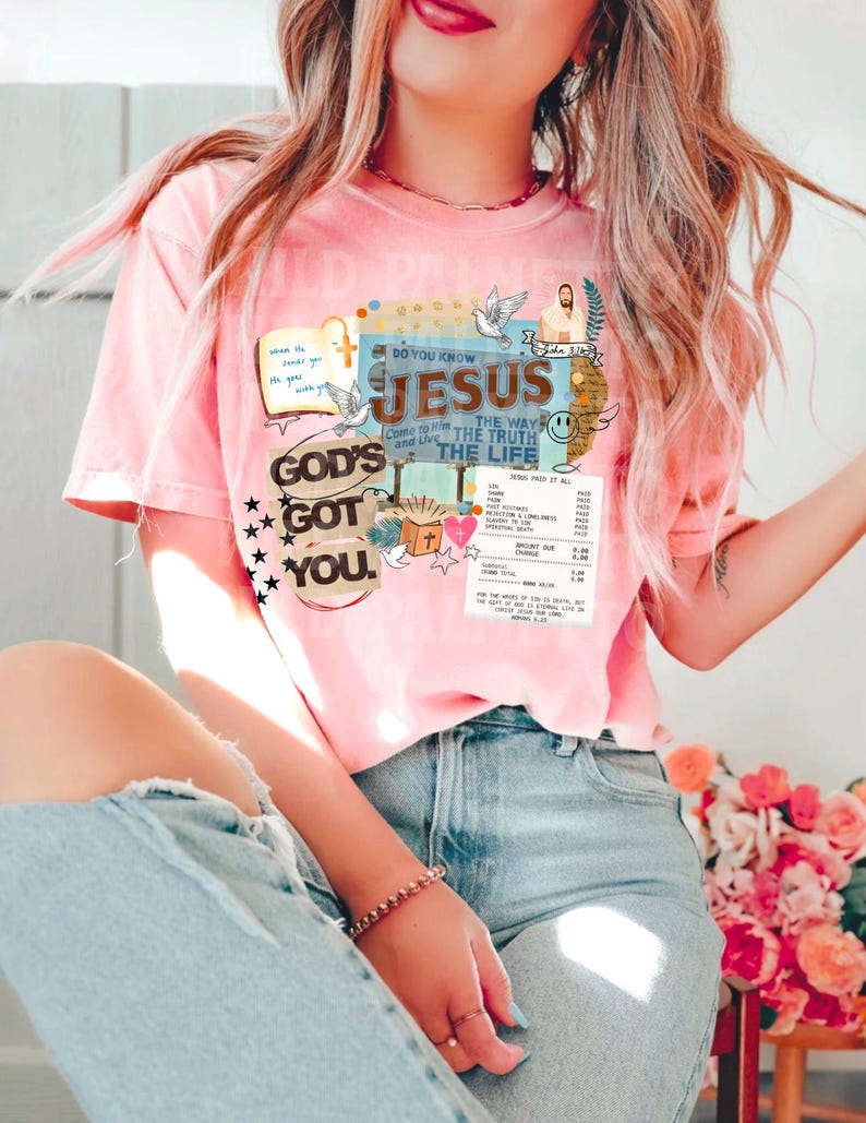 May include: A light pink t-shirt with a graphic design featuring the text "DO YOU KNOW JESUS THE WAY THE TRUTH THE LIFE" and "GOD'S GOT YOU." The design includes doves, a cross, and a depiction of Jesus. The shirt is short-sleeved.
