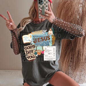 May include: A gray t-shirt with the text "GOD'S GOT YOU" and "DO YOU KNOW JESUS". The shirt has a graphic design with doves, a cross, and the words "THE WAY, THE TRUTH, THE LIFE". A red and white hat that says "TIMELESS" is also visible.