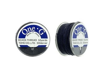 Puede incluir: Dos carretes de hilo para abalorios negro, cada uno con una etiqueta blanca que dice "One 'G' Made in Japan BEADS THREAD 50yards TOHO CO.,LTD. 300denier"