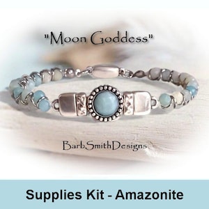 May include: A bracelet with the text "Moon Goddess" features a central round, light blue stone surrounded by a silver-colored frame. The bracelet is made of small, light blue and gray beads. The text "BarbSmithDesigns" is also visible.