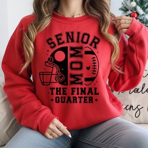 May include: A red sweatshirt with the text "SENIOR MOM THE FINAL QUARTER" in black. The design includes a football helmet and a football. The sweatshirt is a crew neck style.