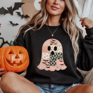 Puede incluir: Sudadera negra con temática de Halloween con un diseño de fantasma de patchwork con murciélagos, calaveras y caras de fantasmas en rosa, negro y blanco. Se sostiene una calabaza naranja tallada.