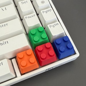 May include: A white computer keyboard featuring standard white keys with black text including Ins, Del, Pause, End, Pgup, and Pgdn, alongside four custom colorful keycaps shaped like building blocks in green, red, blue, and orange colors.