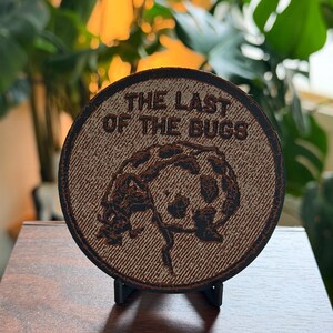 May include: A circular brown patch featuring a textured surface with the text THE LAST OF THE BUGS engraved in dark brown lettering. The center of the patch displays a detailed graphic illustration of a beetle, rendered in dark brown lines.