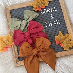 May include: Three fabric hair bows in different colors: sage green, burgundy red, and burnt orange. The bows are arranged on a gray letter board with the text "CORAL & CHAR" in white letters.