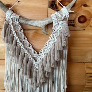 May include: A macrame wall hanging featuring a natural antler base. The design incorporates off-white and tan cotton fibers, with a woven top section and cascading fringe. The piece includes decorative tassels and a neutral color palette.