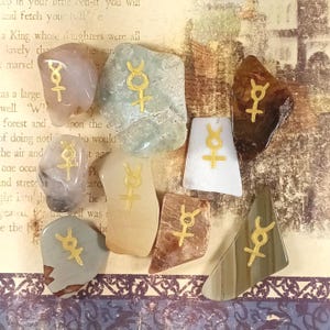 May include: A collection of eight irregularly shaped stones, each featuring a gold-colored symbol. The stones vary in color, including shades of beige, gray, and brown. The background shows a vintage paper with faded text.