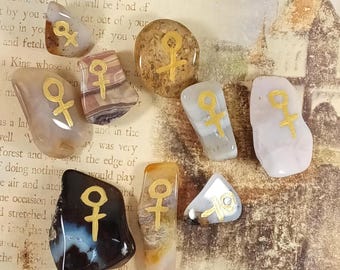 May include: A collection of nine smooth, polished stones in various shades of brown, gray, white, and black. Each stone features a hand-painted gold female gender symbol on its surface, showcasing unique natural patterns and textures.