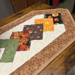 May include: A quilted table runner with a brown, beige, and orange patchwork design. The runner has a scalloped edge and is made of cotton fabric.