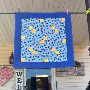 May include: A blue and yellow patchwork quilt with a blue border. The quilt is made up of squares of blue fabric with yellow squares in the center. The quilt is hanging on a clothesline.