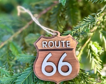 Op de afbeelding: Houten Route 66-bordornament. Het ornament heeft de vorm van een verkeersbord met de woorden "ROUTE" bovenaan en het nummer "66" in grote witte cijfers. Het ornament hangt aan een touwtje van natuurlijk touw en staat tegen een achtergrond van groen gebladerte.