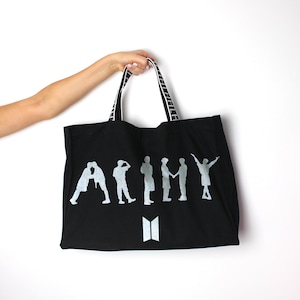 May include: Black canvas tote bag with white striped handles. The bag features a silver silhouette of seven figures with the word "ARMY" printed in silver. The bag also has a silver "BTS" logo.