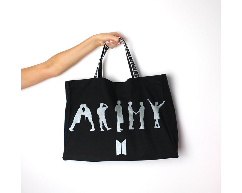 May include: Black canvas tote bag with white striped handles. The bag features a silver silhouette of seven figures with the word "ARMY" printed in silver. The bag also has a silver "BTS" logo.