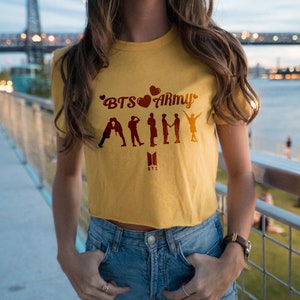 May include: A yellow cropped t-shirt with the text "BTS&hearts;ARMY" and a silhouette of seven figures. The shirt has a small black "BTS" logo below the text.