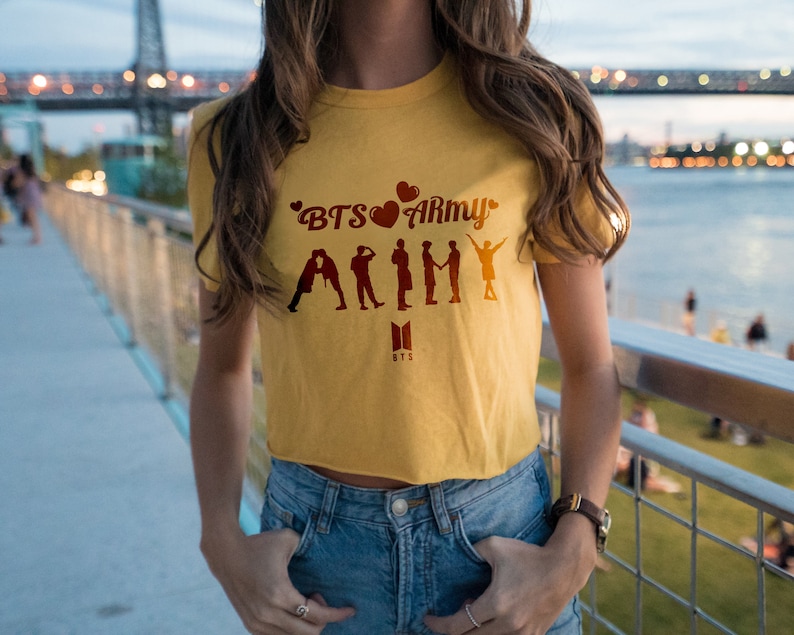 May include: A yellow cropped t-shirt with the text "BTS&hearts;ARMY" and a silhouette of seven figures. The shirt has a small black "BTS" logo below the text.