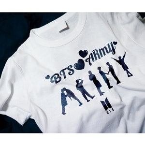 May include: White t-shirt with a black graphic print of the text "BTS Army" with hearts and silhouettes of seven people. The text "BTS" is printed in black below the graphic.