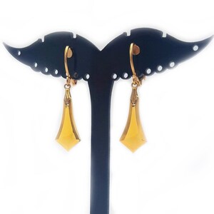 May include: A pair of gold-tone dangle earrings with a geometric design. The earrings are displayed on a black stand.