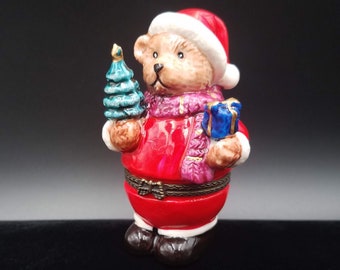 May include: A ceramic Christmas bear figurine wearing a Santa hat and red coat, holding a blue gift box and a small green Christmas tree. The bear has a pink scarf and black boots. The figurine is a hinged box.