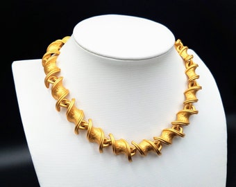 May include: A gold-toned necklace with a textured, spiral design. The necklace is displayed on a white bust against a black background. The necklace's links have a textured surface and a twisted, spiral shape, creating a bold, eye-catching look.