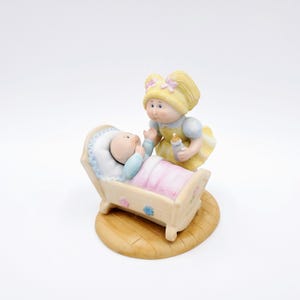 Könnte beinhalten: Porzellanfigur mit einem Baby in einer Wiege und einem Mädchen, das eine Flasche hält. Das Baby ist in eine rosa Decke gewickelt. Das Mädchen hat blondes Haar und trägt ein gelbes Kleid. Der Sockel ist hellbraun.