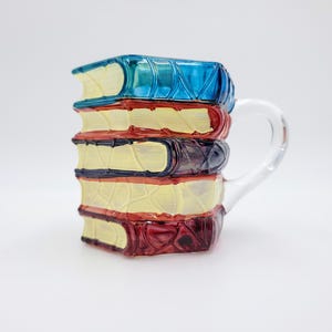 Op de afbeelding: Een keramische mok in de vorm van een stapel van vijf boeken met crèmekleurige pagina's en ruggen in blauw, rood, donkerblauw, geel en bordeauxrood. De mok heeft een helder glazen handvat aan de rechterkant en een textuur op de boekenruggen.