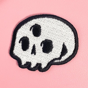 Op de afbeelding: Een witte schedelpatch met zwarte geborduurde details, met drie oogkassen en een gestikte omtrek. De patch is geplaatst tegen een roze achtergrond, met een doorzichtige hoes met een kattenontwerp in de linkerbovenhoek.