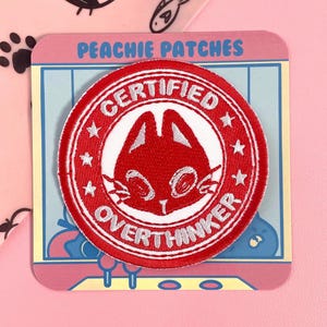 Könnte beinhalten: Ein roter und weißer gestickter Aufnäher mit einem Cartoon-Katzen-Gesicht und dem Text "CERTIFIED OVERTHINKER". Der Aufnäher befindet sich auf einer rosa und blauen Karte mit dem Text "PEACHIE PATCHES". Im Hintergrund sind weitere Katzenmotive zu sehen.