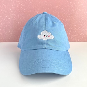 Puede incluir: Una gorra de béisbol azul claro con una nube blanca bordada con una cara sonriente y mejillas rosadas.