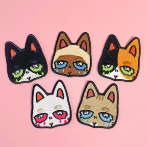 May include: Five embroidered cat face patches in various colours. The cats have sad expressions with tears and green or red accents. The patches are displayed on a pink background.