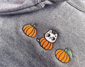 May include: A grey sweatshirt with an embroidered design of a white cat wearing a pumpkin costume. The cat is surrounded by two orange pumpkins.