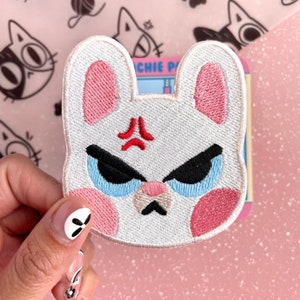 May include: White embroidered iron-on patch of an angry bunny with pink ears, red eyebrows, and blue eyes. The bunny has a frowning mouth and a red angry mark on its forehead.